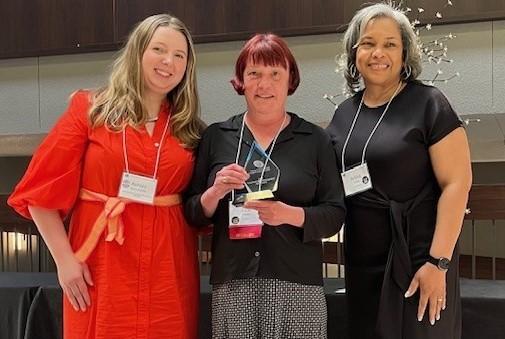 a picture of three people smiling and standing together facing a photographer with the person in the middle holding an award